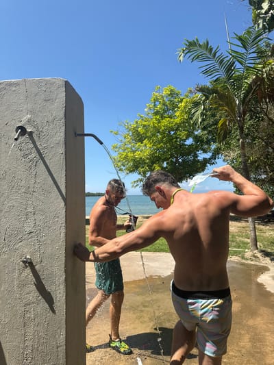 Two adult males are depicted using an outdoor shower facility under clear blue skies in Rio Bueno, Jamaica. The facility consists of a tall, grey concrete pillar from which a metal shower fixture extends, actively dispensing water downwards.
The male on the left, observed partially from his left profile, stands directly under the main shower stream. He wears dark green swim shorts and multi-colored water shoes. A black lanyard is around his neck, and he holds a dark cylindrical object with his right hand. Water is actively streaming over his head and body, splashing onto the paved ground beneath.
The male on the right, viewed from the rear, is positioned slightly to the right of the main shower. He wears pastel-patterned swim shorts with a black waistband and a green and yellow lanyard around his neck. With his left arm raised, he pours water over his head and upper back using a light-colored scoop or container. Water drips from his body onto the wet, light-colored paved surface.
In the background, a calm body of water is visible under bright sunlight, bordered by lush green foliage and distant trees, including a prominent palm tree to the right. The scene is illuminated by strong overhead sunlight, casting distinct shadows.