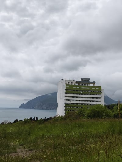 The image shows a tall, white building with green accents located in Ut'os, Ukraine. The building has multiple floors with windows and balconies. It is positioned on a hillside covered with grass and vegetation. In the background, there is a mountain partially obscured by clouds. A body of water is visible in the distance between the mountain and the hillside. The sky is overcast with heavy, gray clouds.