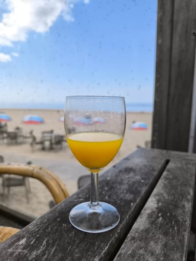 A wine glass filled with yellow juice sits on a weathered wooden surface, likely an outdoor table. In the background is a sandy beach with scattered tables, chairs, and blue-and-red parasols. The sky is blue with scattered clouds. The scene suggests a beachside cafe or restaurant, and the presence of rain drops on the glass suggests a recent shower. A grey wooden wall stands to the right side. The location is likely on a beach somewhere in the Netherlands but it is impossible to confirm that it's Bergen without further information.