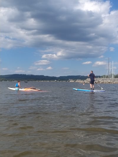 This image depicts a serene scene on the water, likely in or near Radslav, Czechia, given the context provided. In the foreground, the choppy surface of a body of water, possibly a lake or river, dominates the lower half of the frame. The water appears to be a murky brown, with visible ripples and reflections.
To the left of the frame, a person is partially submerged in the water, seated on what appears to be a windsurfing board that has capsized. The person is wearing a blue life vest and appears to be adjusting or righting the equipment. The windsurfing board itself is white with an orange sail attached.
Further to the right, another person stands upright on a paddleboard. This individual is wearing a dark blue t-shirt and black shorts, holding a paddle in their right hand. The paddleboard is light blue with white lettering on its surface.
In the background, a tree-lined shore with a rocky embankment is visible. Beyond the shore, a low hill covered in dense forest stretches across the horizon. The sky above is a vibrant blue, dotted with large, grey, and white clouds, suggesting dynamic weather conditions. In the far distance, several white poles resembling sailboat masts are visible near the shore, hinting at a potential marina or recreational area. The overall impression is one of outdoor recreation on a partly cloudy day.