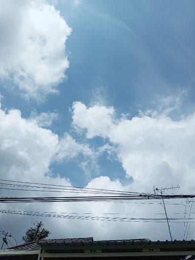 The image depicts an urban sky over Viengthong, Laos, characterized by a light blue expanse with dispersed white cumulus clouds. The lower third of the frame reveals the upper sections of buildings, specifically two adjacent tiled roofs appearing in brown or reddish-brown hues. Multiple dark electrical or communication cables horizontally traverse the middle ground, suspended above the structures. On the right side, a metallic Yagi-style antenna with several cross-elements extends upwards from behind the roofs, intersecting the cable network. On the far left, a partial view of a curved metal structure, possibly part of a satellite dish mount or another antenna, is visible above the roofline. A small cluster of dark green foliage from a tree is discernible behind the roofs in the center-left portion of the image. No individuals, discernible actions, or direct interactions are present.