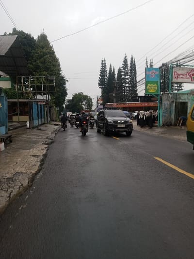 The image depicts a damp urban street scene in Chagzam_Pam, Bhutan, under an overcast sky. The asphalt road, wet from recent precipitation, features a solid yellow line. In the mid-ground, a dark-colored SUV, bearing license plate 'F 1007 FES' and equipped with a roof rack, travels towards the viewer. Behind it, a group of at least four motorcycles, ridden by helmeted individuals, proceeds. On the right-side sidewalk, approximately eight pedestrians, uniformly dressed in dark robes and head coverings, walk away from the camera. Further right, an individual in a blue shirt is observed bent over near a partially visible yellow and green vehicle. The left foreground shows a cracked concrete sidewalk alongside structures with corrugated metal roofs. The background features tall evergreen trees, utility poles, overhead power lines, and commercial signage, including a sign with the text "Rumah."