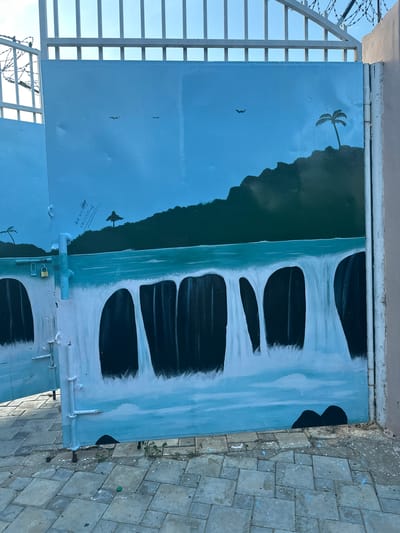 The image shows a mural painted on a metal gate in Batagarawa, Nigeria. The mural depicts a tropical scene with a waterfall cascading down a dark green cliff face into a turquoise body of water. In the background, a silhouette of a palm tree stands against a clear blue sky. Two small birds are painted in the sky. There is also an inscription on the left side of the mural, which appears to be a signature and a date. The gate itself is painted light blue and is slightly weathered. A padlock is visible on the left side of the gate. The ground in front of the gate is paved with rectangular stone tiles and shows some scattered paint splatters. A white metal fence with barbed wire is visible above the gate.