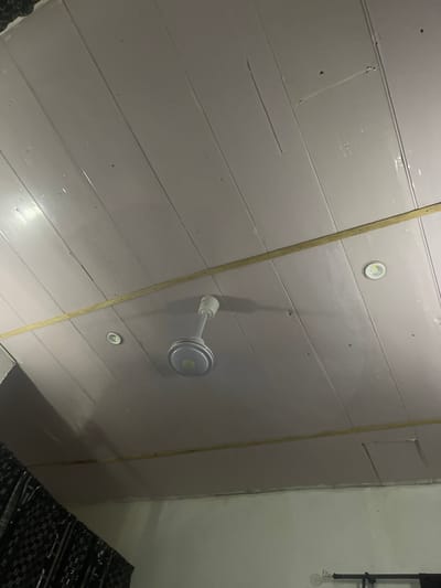 The image displays a low-angle view of a room's ceiling, which is constructed from horizontal wooden planks painted a light, matte gray. A thin, light-colored wooden strip runs diagonally across the ceiling, attached with visible nails. Several circular LED ceiling lights are embedded in the planks. In the upper right quadrant of the image, a white ceiling fan with metallic blades is partially visible, attached to the ceiling. To the right of the ceiling fan, a black metal pipe structure with a hanging light fixture is attached to what appears to be a wall or a raised platform. The lower right corner of the image shows a dark-colored, textured material, possibly a rug or mat with a repeating pattern. The lighting in the room suggests it might be nighttime or a dimly lit indoor environment, with the ceiling lights providing illumination. The overall scene depicts the interior of a room with a focus on its architectural elements and lighting fixtures. The location context provided is Pantinapu, Nigeria, but there are no specific visual cues within the image to confirm this location. No people are visible in the image. No text is discernible.