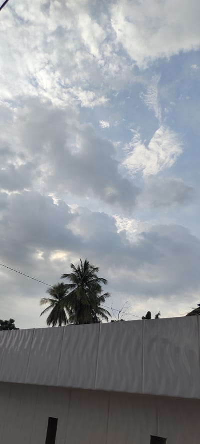 The image is a vertical, outdoor shot taken at ground level, looking upwards. The foreground is dominated by a white, textured wall with visible seams running vertically. Above the wall, two coconut trees with lush green fronds are silhouetted against a cloudy sky. The sky is a mix of light blue and grey clouds, with patches of brighter sky visible through breaks in the clouds. A thin black wire stretches horizontally across the frame above the trees. The scene appears to be a view of the sky and vegetation from behind a wall, possibly in a residential or urban area of Meunasahaluedrienlhoksukon Barat, Indonesia. No people are visible in the frame.