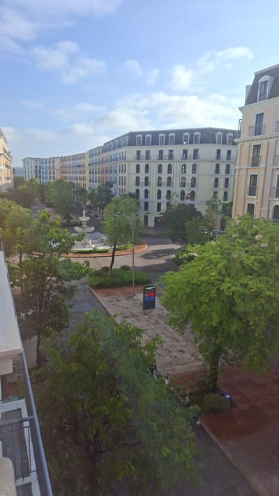 The image shows a view from an elevated position, possibly a balcony, overlooking a street in Phú Quốc Special Administrative Region, Vietnam. A large, beige-colored building with multiple floors and numerous windows is prominent on the left side of the image. The building appears to be a residential or commercial complex. A street lined with trees and greenery runs through the center of the frame, leading away from the viewer. There is a fountain visible along the street, closer to the bottom of the image. A sign with a digital display is located along the right side of the street. The sky is visible in the upper left portion of the image and is a pale blue with some clouds. The overall scene suggests a residential or mixed-use area in the city.