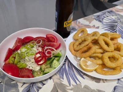 A meal is laid out on a table with a floral patterned tablecloth. On the left, a white bowl contains a salad with chopped tomatoes, cucumbers, green peppers, red peppers, onions, and a creamy dressing, sprinkled with herbs. To the right of the salad bowl is a white plate piled with golden-brown fried calamari rings, with a slice of lemon on the side. Behind the food, a dark brown bottle of Ottakringer beer stands upright. The scene is likely indoors, possibly a dining area in Bruck an der Leitha, Austria, given the mention of the beer brand which is Austrian. The lighting appears to be from an artificial source. No people are visible in the image.