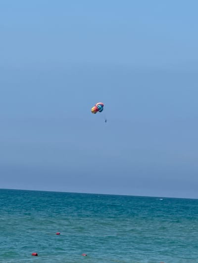 The image shows a bright, clear day with a vast expanse of light blue sky dominating the frame. In the upper left portion of the sky, a colorful parachute is visible, appearing to be engaged in a parasailing activity. The parachute is a vibrant mix of red, yellow, and blue hues, suggesting it's a recreational activity. Slightly above and to the right of the parachute, a small, dark silhouette of a person or object is also present in the sky, possibly a bird or another, smaller kite.

The right side of the image is filled with the deep blue-green waters of the sea. The water has a textured appearance, indicating movement and ripples on the surface, likely due to a gentle breeze. Scattered across the water are several small, red buoys, marking a course or designated area within the sea. The overall impression is one of a sunny day at a beach or coastal resort, with recreational activities taking place in the sky and on the water. The clarity of the sky and the vibrant colors suggest it is daytime, likely midday or early afternoon, under fair weather conditions. The presence of the parachute and buoys points to a location where water sports and leisure activities are common, such as a beach resort area. No text is visible in the image.