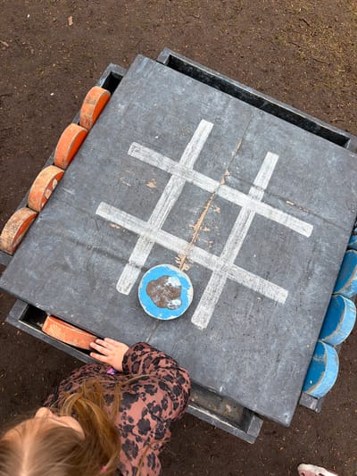 The image captures an outdoor tic-tac-toe game in what appears to be a recreational space. The game is played on a square, gray-surfaced structure marked with white lines creating the grid. One game piece, a blue circle with a brown design, is placed on the board.
The game board sits on a wooden pallet-like frame. Wooden circles painted blue and orange are tucked into the frame, likely serving as game pieces. A child wearing a brown spotted jacket is partially visible, with her hand near the edge of the game and a glimpse of her face and blonde hair. The area surrounding the game board is covered in dirt and sparse vegetation. The setting suggests a playground or park. The location given is in the city of Knutsford, United Kingdom.