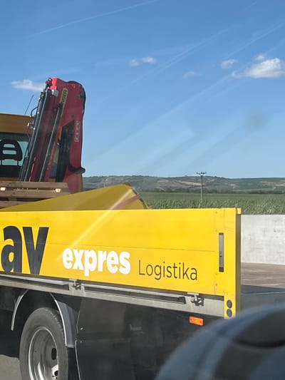 A yellow truck with "av expres Logistika" written on its side is parked on a road in Rakvice, Czechia. The truck has a crane on its back, which is red and has "KUN" written on it. The truck is parked next to a field of green crops and a hill in the background. The sky is blue with some white clouds.