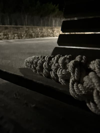 An outdoor night scene in Tyulenovo, Bulgaria, shows a thick, braided rope lying horizontally across a dark wooden surface. The rope is textured and appears to be made of natural fibers. To the left of the rope, a paved surface is visible, with a low stone wall and trees in the background, illuminated by ambient light. To the right of the rope, the dark, slatted structure of a bench or seating area is present. The lighting is dim, with shadows obscuring details in the foreground and background, creating a moody and somewhat abstract composition. No people are present in the image.