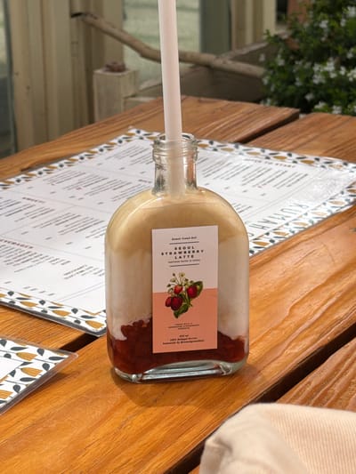 The image captures a strawberry latte drink in a glass bottle on a table at Rumah Gemak Bali, a restaurant in Candikuning, Indonesia. The table has a printed menu with a floral design. A white straw sticks out of the bottle. A portion of a person's white shirt can be seen in the top-right corner. The table is made of light brown wood. A plant is visible in the background near a window.