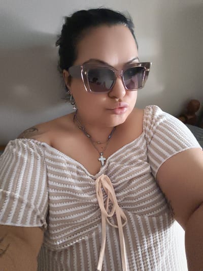 The image shows a close-up, medium shot of a woman with dark hair pulled back, wearing large, square, translucent purple sunglasses. She has full lips and is looking directly at the camera. She is wearing a cream and beige striped off-the-shoulder top with a drawstring detail at the neckline. Around her neck, she wears two necklaces; one with small beads and another with a silver cross pendant. She has small hoop earrings and a tattoo visible on her left shoulder. The background is a plain, lightly textured wall, with a portion of a bed and a stuffed animal visible on the right. The lighting suggests an indoor setting, possibly a bedroom. The image does not provide any information about the location within the city of Komotini, Greece.