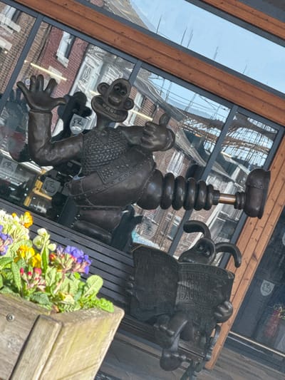 The image displays a bronze sculpture located on Corporation Street, Preston, United Kingdom. In the central area, a bronze statue of Wallace is depicted standing, wearing the "Techno Trousers" from "The Wrong Trousers." Wallace's left hand is raised in an open-palm gesture, while his right hand forms a thumbs-up. To the lower right of Wallace, a bronze statue of Gromit is seated on a bench, appearing to read a newspaper titled "The Feathers McGraw Back Inside." Both figures are positioned in front of a building featuring large, reflective glass windows that mirror surrounding red-brick architecture, scaffolding, and a bright sky, indicating an outdoor urban environment. A wooden planter filled with various colorful flowers, including shades of purple, yellow, and white, occupies the blurred foreground on the left.