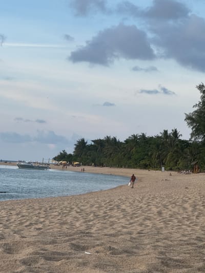 The image depicts a beach scene in Baan Wang Ta Kien, Thailand. A coastline of palm trees separates the sand from the sky. A person wearing a red shirt is walking along the beach with an object in their hands. The sand is tan, and the water is blue-grey. The sky is overcast with cloudy skies.