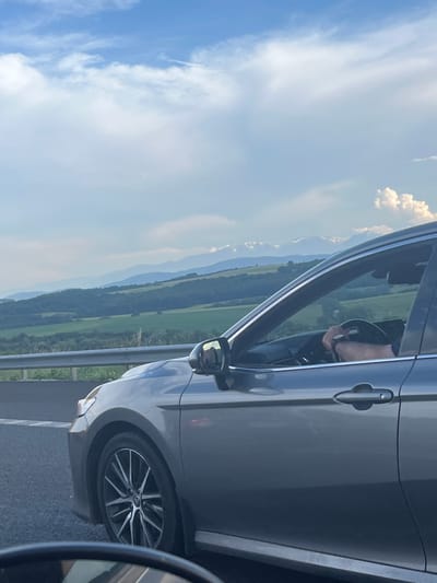 The image shows a person driving a gray car on a road, with a scenic landscape in the background. The sky is a bright blue with scattered white clouds, suggesting a clear and sunny day. The landscape features rolling green hills and distant mountains, creating a sense of depth and natural beauty. The car's interior is partially visible, showing the steering wheel and the driver's arm. The side mirror of the car also reflects the surrounding environment, offering another glimpse of the scenery. The image appears to be taken during the daytime, likely late morning or early afternoon. The overall mood is peaceful and serene, capturing a moment of travel through a picturesque rural area. The location is specified as Vakarel, Bulgaria, which adds context to the mountainous terrain and greenery. There is no visible text in the image.