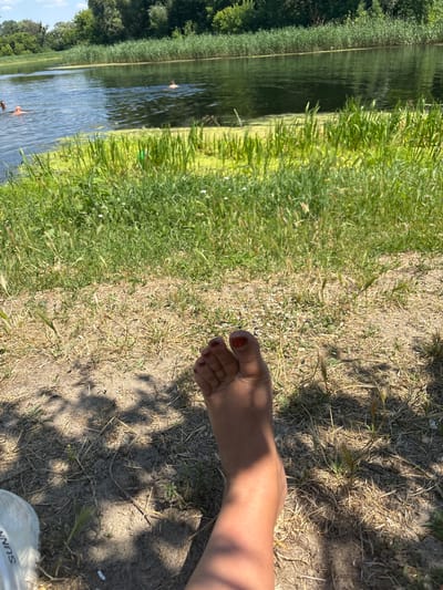 A person's bare foot with red nail polish is in the foreground on the right side of the image. The foot is on dry, sandy ground with scattered grass and straw. There are shadows cast on the ground, suggesting it is a sunny day. To the left of the foot, there is a grassy area, and beyond that, a body of water. The water is calm and reflects the surrounding greenery and sky. Along the edge of the water, tall reeds or grass are visible. In the water, there appear to be at least two people swimming. The overall scene depicts a relaxed outdoor setting, likely a lake or river on a warm day. The image captures a moment of leisure and summer activity.