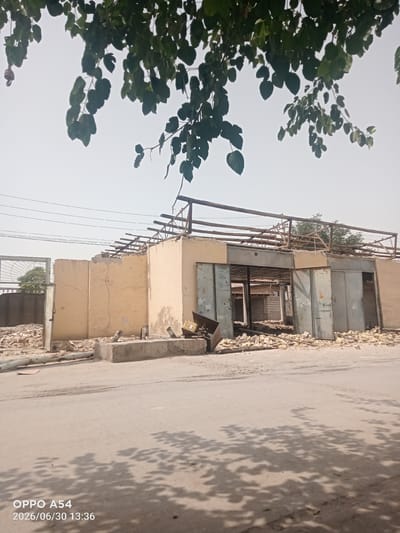 The image shows a partially demolished building with its roof structure exposed. Rubble and debris are scattered around the site, indicating ongoing demolition work. The building appears to be made of concrete blocks with a metal frame supporting the remaining roof. There are large metal doors on one section of the building. Overhead, tree branches with green leaves obscure the sky. In the foreground, a paved surface, likely a road, shows shadows cast by the leaves. A timestamp in the bottom left corner reads "OPPO A54 2026/06/30 13:36". The overall scene suggests a site undergoing reconstruction or demolition in an urban or semi-urban environment.