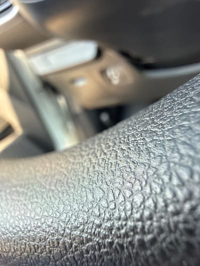 The image is a close-up, out-of-focus shot of a textured, dark surface, likely part of a car's interior, such as a steering wheel or dashboard. The texture appears to be a grainy, dimpled pattern. In the blurred background, lighter colored objects and surfaces are visible, possibly indicating other parts of the car's interior or reflections from the outside. The lighting suggests an indoor or shaded environment, possibly during the day due to the ambient light. There are no people or specific landmarks visible that would allow for identification of a location in Soroca, Moldova.