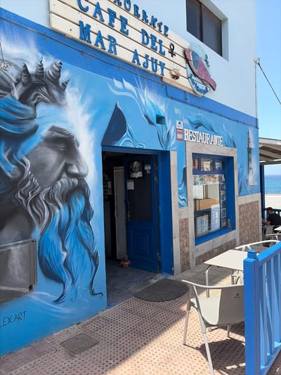 A vibrant, art-adorned cafe or restaurant building stands out with a predominantly blue and white facade. The exterior is covered in elaborate murals featuring crashing waves, a stylized lighthouse, and a large depiction of a mythical figure, possibly a sea goddess or siren, with flowing blue hair or drapery.
Outdoor seating with white tables and chairs is arranged on a tiled patio, illuminated by bright sunlight, casting distinct shadows. In the upper background, beyond the building and seating area, a sandy beach and the blue sea are visible, with a person standing near the water.
A prominent wooden sign on the left side of the building reads "CAFE DEL MAR AQUI" (Cafe of the Sea Here), with blue letters affixed to light wooden planks. Above this sign, a red-hued fish is mounted on a circular base. Another sign, above a display window, spells out "RESTAURANTE" in white letters. The display window itself showcases various food items.
Notable details include an alarm sticker with "Alarma Grimalt" and a phone number next to the "RESTAURANTE" sign. The signature "ALEX.ART" is visible on the lower right portion of the mural, indicating the artist. The sunny weather and clear blue sky suggest it's daytime, possibly midday or early afternoon. The overall scene conveys a cheerful, seaside atmosphere in Pájara, Spain.