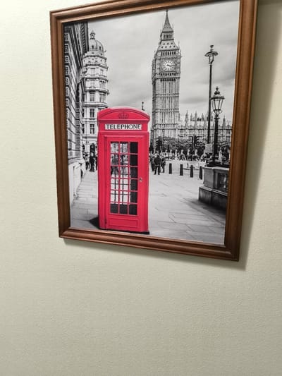 The image displays a framed artwork depicting a black and white scene of a London street with a vibrant red telephone booth in the foreground. Behind the telephone booth, the iconic Big Ben clock tower and other London architecture are visible. The artwork is hung on a textured, cream-colored wall. The framing is made of wood with a dark, reddish-brown finish. The image is taken from an angle, showing the wall and a portion of the ceiling. The scene is static and captured indoors. There is no indication that this image is located in Nowogard, Poland.