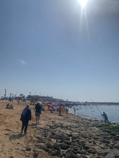 The image depicts a crowded beach scene on a sunny day in Harhoura, Morocco. The foreground is dominated by a sandy shoreline with rocky outcrops. Two individuals are walking away from the camera on the sand in the lower left quadrant. In the mid-ground, numerous people are scattered across the beach, many sitting under colorful umbrellas. The water is populated with many swimmers. A man is actively fishing with a rod and reel on the rocky edge of the water in the right mid-ground. In the background, several buildings and palm trees line the coast. The sky is clear blue with the sun prominently visible in the upper right, casting a bright glare. A kite is visible flying in the sky.