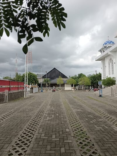 The image depicts an outdoor scene on a cloudy day, likely in the late morning or early afternoon. The foreground is dominated by a paved area made of interlocking concrete blocks, with diagonal strips of open diamond-shaped holes filled with grass running through it, suggesting a parking area or walkway.

In the midground, several buildings are visible. To the right, a white building with a blue, patterned dome, characteristic of a mosque, stands prominently. It features arched windows with intricate lattice work. To the left and center, there are other buildings, one with a dark, angled roof, and further back, residential-looking structures. Behind these buildings, several tall red and white communication towers rise against the overcast sky. Trees with green foliage are scattered throughout the scene, adding to the natural elements.

There are a few people visible in the image, but they are small and not the primary focus. One person is riding a red motorcycle in the middle ground, near the buildings. Others appear to be seated or standing near the mosque's entrance.

The weather appears to be overcast, with heavy clouds obscuring the sun, creating a diffused light and a somewhat muted color palette. The overall impression is of a tranquil, possibly slightly humid, day. There is a signpost with blue text and some white lettering on it near the mosque's entrance, but the text is too small to be deciphered. The setting appears to be a community area, possibly a mosque complex, given the presence of the mosque and the surrounding buildings.