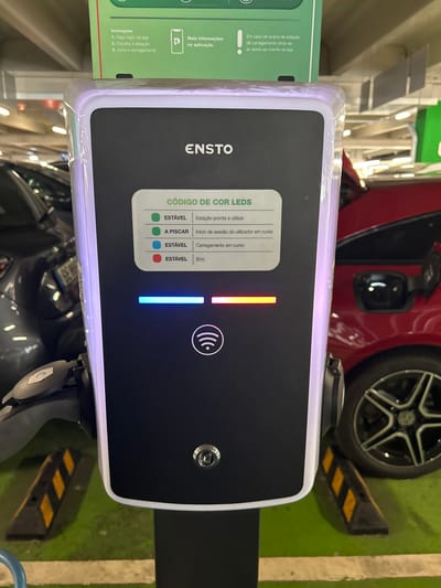 A full-shot, eye-level, outdoor, eye-level, vertical photograph captures an Ensto electric vehicle charging station in a parking garage. The charging station is positioned centrally and illuminated with white and purple light around its edges. It features a black screen displaying information in Portuguese about LED indicators, with options for "Estável" (Stable) and "A piscar" (Blinking). Below the screen are illuminated LED bars: a blue one on the left, a yellow one in the middle, and a red one on the right. A Wi-Fi symbol is displayed beneath the LED bars.
To the left of the charging station, a portion of a dark-colored car is visible, and to the right, a red car is parked. The ground is covered in green asphalt with yellow and black striped parking guides. The scene appears to be in the parking area of a shopping center, potentially in Senhora da Hora, Portugal, as indicated by the presence of the Ensto charging station, a brand commonly found in Europe. There are no people present in the frame. The overall lighting suggests it is daytime, with artificial lights from the parking garage also contributing.