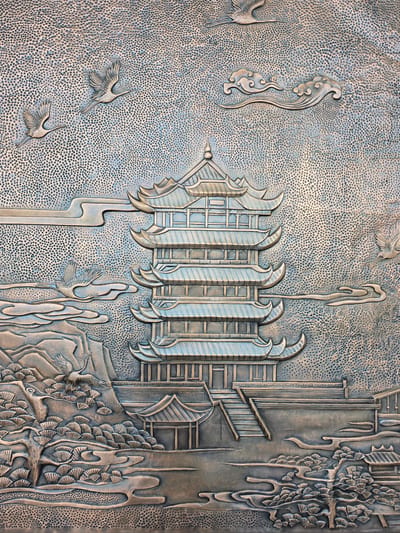 This is a detailed relief sculpture depicting a traditional Chinese landscape with architectural elements and wildlife. The central focus is a multi-tiered pagoda with distinct curved eaves, suggesting classical East Asian architecture. Flanking the pagoda are stylized clouds, and in the foreground, there are elements of nature including pine trees with fan-shaped needles and mountainous terrain. Several cranes are depicted in flight throughout the scene, symbolizing longevity and good fortune. The background of the sculpture is textured with numerous small, raised dots, creating a sense of depth and atmosphere. The overall color palette is metallic, with shades of silver and blue, implying the material is likely bronze or a similar metal. There are no people or contemporary elements visible. The image does not provide any information about its location in the city of Wuhan, China.