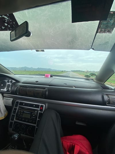 The image depicts the interior of a car, likely in motion, with a view of a road and landscape through the windshield. A dark grey dashboard dominates the right side of the frame, featuring air vents, a radio console, and what appears to be a gear stick. The passenger is wearing a dark pinstripe suit. A red bag is visible as well. The windshield shows a road stretching into the distance, flanked by green vegetation and hills, under a hazy sky. Traffic stickers are affixed to the upper part of the windshield. There is no indication of Assob Hausa, Nigeria, in the image.
