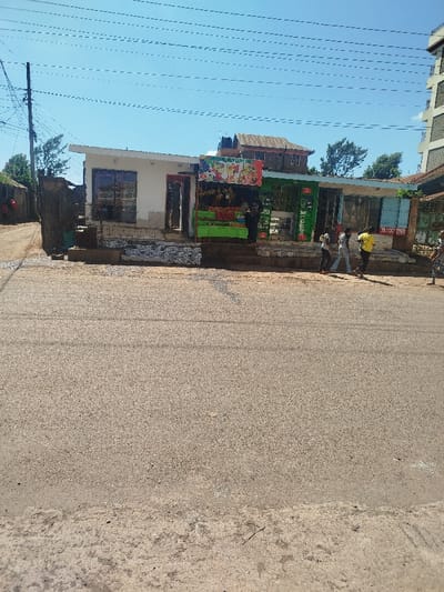 The image captures a street scene in Kihara ward, Kenya, under a clear blue sky. The foreground is dominated by a grey asphalt road. Above the road, a raised sidewalk fronts a series of commercial structures.

From left to right, the establishments include a white-painted building with two dark-framed windows and a reddish-brown entrance door. Adjacent to this is an open-fronted fruit and vegetable stall, displaying produce such as bananas, apples, oranges, and watermelon on a green-covered table. A multi-colored sign depicting various fruits hangs above this stall. To its right are additional shops, one featuring "M-PESA" branding with green and light blue corrugated metal siding.

Several individuals are present on the sidewalk. One person in dark clothing stands behind the fruit stall's display. To the right of the fruit stall, three individuals walk from left to right: one in a light grey t-shirt, one in a white t-shirt, and one in a yellow t-shirt, all wearing dark pants. A fourth person, partially visible, stands on the far right.

In the mid-ground and background, a brown-roofed building with a black water tank on its roof is visible. Further to the right, a taller, multi-story building with a light-colored facade rises. Overhead, multiple utility lines traverse the upper portion of the frame, supported by a utility pole positioned on the far left. A dirt path branches off to the left of the white-painted building.