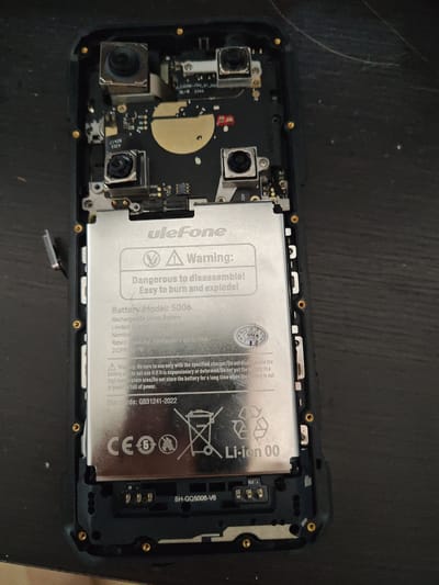 The image displays the internal components of a disassembled Ulefone model 5006 smartphone. The main subject is the removable, silver-colored Li-ion battery, which has a capacity of 7800mAh. The battery is labeled with warnings regarding disassembly and safe usage, including a "Dangerous to disassemble! Easy to burn and explode!" caution. Text on the battery also indicates it is a Rechargeable Li-ion Battery with a nominal voltage of 3.8V. Below the battery, a black circuit board is visible with various connectors and markings, including "SH-GQ5006-V6" and "CC2". Two camera modules are also present on the circuit board, situated at the top of the phone's internal layout. The overall environment appears to be a dark, flat surface, possibly a desk or table, with some faint scratches or dust visible. There are no people or any indications of the city of Mělník, Czechia, present in the image.