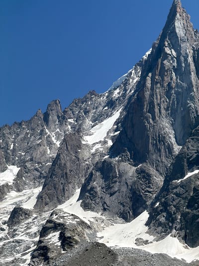 This media file is a close-up, angled shot of a dramatic, rocky mountain face with patches of snow. The mountain is sheer and imposing, with jagged textures and deep crevices that suggest a harsh and rugged environment. The snow clings to the rock in irregular streaks and patches, highlighting the contours of the terrain and indicating a high-altitude location. The sky is a clear, vibrant blue, suggesting fair weather and possibly a bright, sunny day. The lighting indicates that the sun is high, casting sharp shadows that emphasize the mountainous topography. The composition is dominated by the dark, textured rock and the stark white of the snow, contrasted against the expansive blue of the sky. The absence of any human figures or discernible man-made objects places the scene firmly in a natural, wild setting. The overall impression is one of immense scale, raw natural beauty, and the powerful forces of geology. The location context provided, Chamonix-Mont-Blanc, France, further reinforces the interpretation of this being a view of the French Alps, a region renowned for its majestic peaks and challenging terrain.