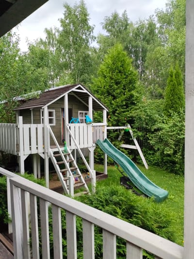 The image shows a children's playhouse with a slide and swing set in a backyard, located in Ермолино-2 Снт, Russia. The playhouse is elevated on stilts and has a wooden structure with a white railing and stairs leading up to it. A green plastic slide extends from the platform of the playhouse to the grassy ground. A swing set frame is visible behind the playhouse. The backyard is surrounded by lush green trees and bushes. There are no people visible in the image. The overall scene depicts a typical suburban backyard setting designed for children's recreation.