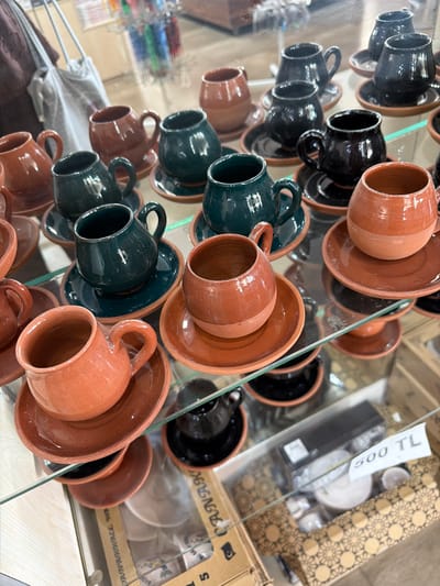 The image shows a close-up, eye-level view of shelves in a shop, likely in Acıpayam, Turkey, displaying numerous sets of small, ceramic cups and saucers. The cups appear to be traditional Turkish coffee cups, with a reddish-brown unglazed bottom half and a glazed top half in colors such as dark green, black, and reddish-brown. The saucers are a matching reddish-brown. Many of the cups are filled with light, and some are stacked. In the background, a grey fabric tote bag is visible hanging, along with other merchandise which is out of focus. A price tag reading "500 TL" is also visible in the lower right corner, indicating the currency of Turkey. The overall impression is one of a retail display of traditional artisan crafts.