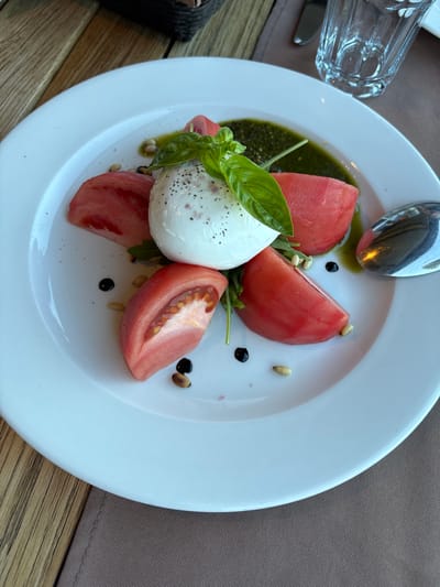 A close-up shot features a Caprese salad on a white plate, positioned on a brown tablecloth on a wooden table. The salad consists of a ball of fresh mozzarella, seasoned with black pepper, topped with basil leaves. It is surrounded by wedges of ripe tomatoes. Drizzled over the dish are balsamic glaze and a vibrant green pesto sauce, with scattered pine nuts adding texture. A silver spoon rests on the right side of the plate. In the background, a clear glass with water and part of a woven basket are visible. The setting appears to be a restaurant in Topola, Bulgaria, suggesting an outdoor or indoor dining area with natural light.