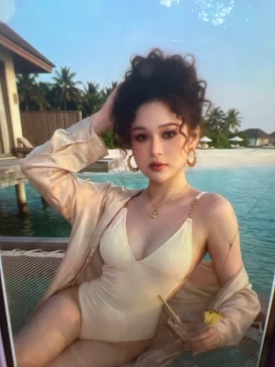 The image shows a young woman in a cream-colored swimsuit and a light-colored open robe, seated on a textured surface next to a body of water. She has dark, curly hair pulled up and is wearing large hoop earrings and a delicate necklace. In her right hand, she holds a drink with a straw and a slice of pineapple. Behind her, palm trees and small buildings are visible, suggesting a tropical or subtropical location. The water appears to be clear and blue. The scene is lit by natural light, likely from the sun. There is no indication of Kara-Dıykan, Kyrgyzstan in the image.