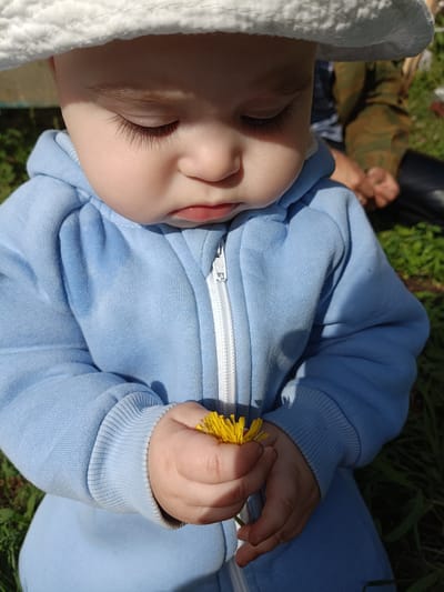 A baby wearing a white hat and a light blue hooded outfit is holding a yellow dandelion. The baby's eyes are closed and the baby is looking down at the flower. The baby is outside in a grassy area. The location, Усть-Тунтор, Russia, cannot be determined from the image.