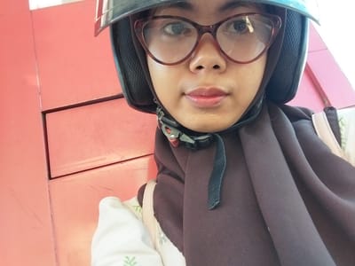 A close-up selfie shows a young person wearing a black helmet and glasses, posing in front of a coral-colored wall. The subject is looking directly at the camera with a neutral expression. They are wearing a brown hijab that covers their head and neck, and a white shirt with a subtle green leaf pattern is visible underneath. The helmet has a chin strap with a buckle. The background appears to be an indoor or sheltered outdoor area, possibly a garage or a covered walkway, with the coral wall showing some scratches and faint marks. The lighting suggests it could be daytime. The setting might be Macege, Indonesia, as indicated. No text is visible in the image.