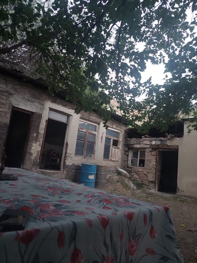 The image depicts the exterior of a weathered, abandoned building, likely in the town of Areni, Armenia. Overhanging tree branches with green leaves partially obscure the view, suggesting an outdoor setting. In the foreground, a table is covered with a patterned tablecloth featuring red and grey floral designs. To the right of the table, a blue barrel is visible, standing on the ground. The building itself appears to be constructed of stone and plaster, with visible signs of decay, including peeling paint and cracked walls. Several windows are present, some with intact panes and others broken or missing. One doorway is open, revealing a dark interior. The ground surrounding the building is unpaved, with scattered debris and dry vegetation. The overall atmosphere is one of neglect and disrepair, hinting at a historical or rural location.