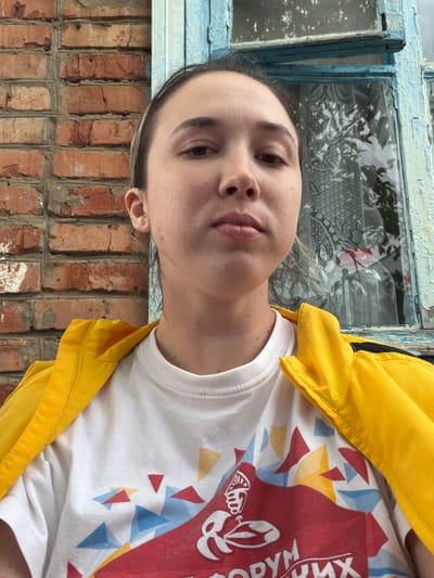 A young woman with light brown hair pulled back is looking directly at the camera. She is wearing a white t-shirt with a red and blue abstract design and a bright yellow zippered jacket. The background consists of a red brick wall to her left and a distressed blue wooden window frame with a sheer white curtain to her right. The woman appears to be outdoors, possibly on a porch or near a building in the city of Komsomolskiy, Russia. The lighting suggests it is daytime.