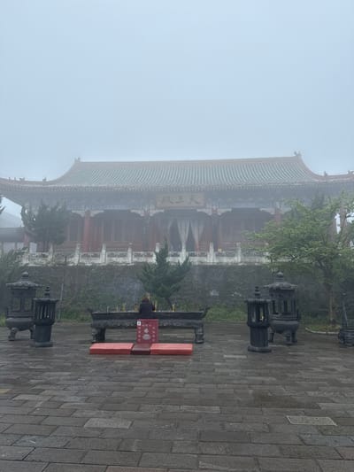 The image depicts a building, partially obscured by fog, in the Yongding District, China. The building has a traditional Chinese architectural style with a multi-tiered roof and intricate window designs. A person stands in front of a red rectangular structure with Chinese characters, possibly a donation box or offering table. Incense sticks are visible near the person. In the foreground and background are stone pavers and ornamental lanterns. The environment is overcast, with heavy fog impacting visibility.