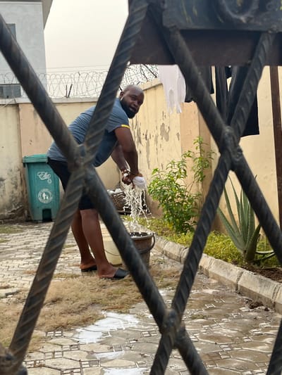 The image captures an outdoor scene, likely a yard or pathway, viewed through the blurry, dark, twisted bars of a metal gate or fence in the foreground. The primary subject is a bald, bearded man, wearing a blue short-sleeved shirt with a yellow stripe on the shoulder and dark shorts. He is barefoot, though a black slipper is partially on one foot.
The man is actively wringing out a white cloth or towel over a dark bucket, causing water to splash outward. The ground around him is paved with patterned tiles, which appear wet in several spots. Patches of green grass and some plants, including a large agave, are visible around the paved area.
In the mid-ground, a green plastic waste bin is situated, featuring a circular white logo. The logo depicts hands dropping waste into a bin and contains visible text, including "CLEAN ENVIRONMENT" and "KEEP." The background reveals a building wall and a fence topped with coiled barbed wire, suggesting a secure or residential area in Dushepe, Nigeria. The lighting is bright and even, indicating it is daytime. The man's expression appears neutral, focused on the task of wringing out the cloth.