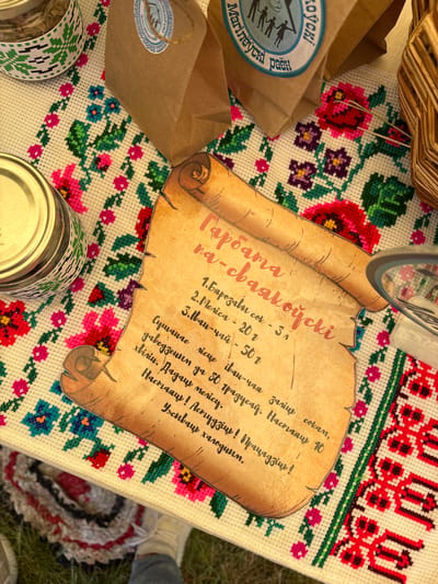 The image shows a close-up, overhead view of a table covered with a white, embroidered cloth featuring floral patterns in red, pink, green, and blue. On the cloth, a rolled-up piece of parchment with handwritten text in red ink is prominent. The parchment appears to be a recipe or instructions, listing ingredients and quantities such as "Барозавы сок - 3л" (Birch sap - 3l), "Mjerica - 20 г" (Mjerica - 20g), and "Іван-чай - 50 г" (Ivan-tea - 50g). Below these, there's further text that reads, "Сушаная гiсце, завядзiшін да 50 градусаў. Наспаддаць 10 хвiлін. Дадаць мёду. Наставдаць! Прагаладзіць!" (Dried herbs, wilted to 50 degrees. Infuse for 10 minutes. Add honey. Steep! Strain!).
To the left of the parchment are two glass jars with metal lids, one containing dried herbs or spices. To the right of the parchment, a woven basket is visible. In the background, two brown paper bags with circular blue and white stickers are partially visible. The stickers have text and an illustration of people. The overall scene suggests an outdoor market stall or a rustic setting. The location is identified as Вяйнянскі сельскі Савет, Belarus.