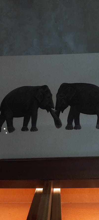 The image shows a silhouette painting of two elephants facing each other, with their trunks intertwined. The elephants are dark gray and are depicted against a lighter gray background. The painting appears to be framed by dark wood at the bottom, with a warm orange light emanating from below the frame, suggesting it is displayed in a well-lit area or is part of a display. The background above the painting is a dark, textured gray, possibly a wall. The setting is identified as Sampora, Indonesia, though no specific landmarks or contextual details about the city are visible in the image itself. The overall impression is of an artistic representation of elephants, potentially within a museum or gallery setting.