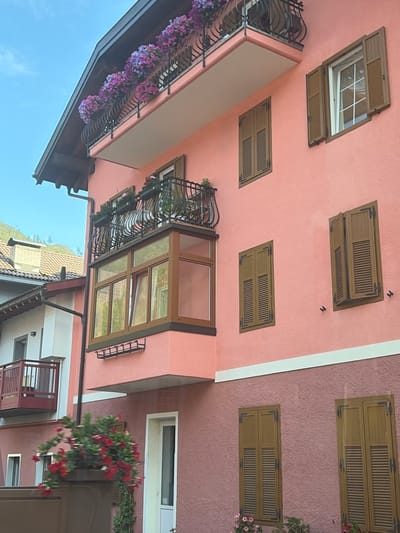 A close-up, side-angle view of a multi-story building in Predazzo, Italy, displays a vibrant pink facade adorned with numerous wooden shutters. The building features multiple balconies, one with a black wrought-iron railing overflowing with pink and purple flowers. Another, smaller balcony with a red railing is visible on an adjacent section of the building. A prominent bay window with wooden frames interrupts the facade. The sky above is clear and blue, and a hint of a mountain landscape can be seen in the background. A planter box with red flowers is positioned in the foreground.