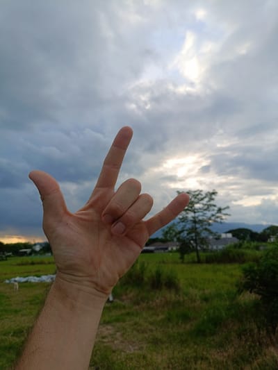 A close-up shot focuses on a person's right hand forming the "I love you" sign, with the thumb and index finger extended and the ring and pinky fingers bent. The hand is held against a backdrop of a cloudy sky and a rural landscape. In the background, a grassy field stretches towards a line of trees and buildings, with mountains visible in the distance. The sky is filled with dramatic clouds, with patches of sunlight breaking through. The overall impression is one of a serene, outdoor setting. The location is identified as Ban Patan, Thailand.