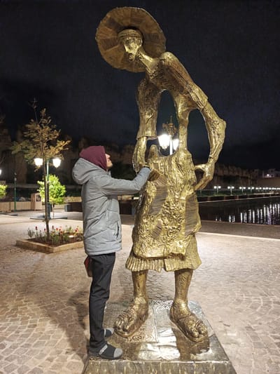 At night in the city of Al Machouar – Stinia, Morocco, a man wearing a grey jacket with a maroon hood, dark pants, and grey casual shoes stands on a patterned pavement beside a large golden statue of a man wearing a large hat. The statue holds an ornate streetlamp fixture in its arms. The man in the jacket is touching the statue with his right hand. In the background are trees, other streetlamps, and a body of water with lights reflected in it. A wall and buildings are visible beyond the water. The sky is dark.
