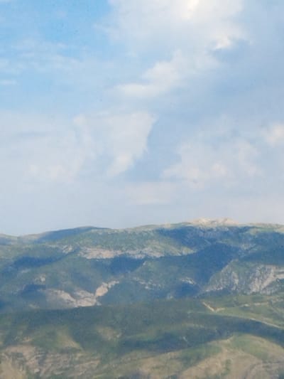 The image depicts a panoramic view of rolling, green-covered mountains under a bright blue sky with scattered white clouds. The landscape is characterized by layers of undulating hills and peaks, with visible shadows indicating sunlight. The vegetation appears dense and varied in color, ranging from deep green to lighter shades. There are no visible man-made structures or people in the frame. The overall impression is one of a natural, undeveloped mountainous terrain, likely an outdoor scene. The specific location in Ак-Дөбө, Kyrgyzstan, is implied by the prompt but not visually identifiable within the image itself.