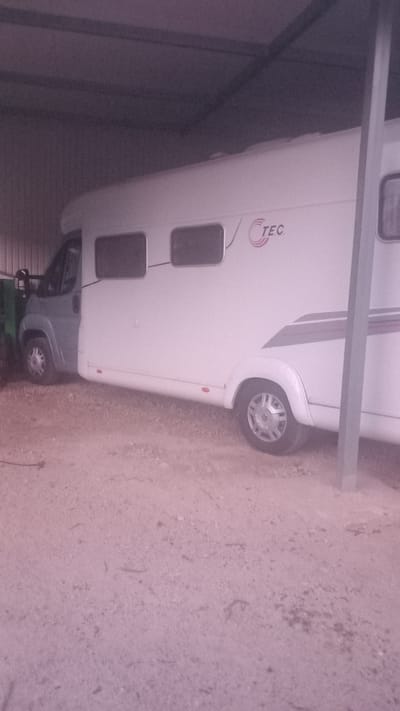 The image shows a white motorhome parked inside a covered structure, possibly a carport or garage. The motorhome is white with gray accents and has several windows visible. A logo with the letters "TEC" is present on the side of the motorhome. The ground is covered in gravel or sand. Another vehicle, partially visible in gray, is parked in front of the motorhome. The lighting suggests it could be daytime, but the setting is indoors or under cover, so external weather conditions are not discernible. There are no people visible in the image. The overall impression is one of storage or parking. The location context provided is Oliveira do Bairro, Portugal, which adds geographical information to the scene.